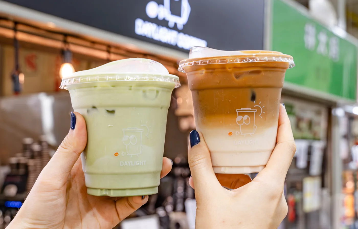 An iced oat white and an iced matcha latte in Daylight Coffee cups, held up in front of the stall at Amoy Street Food Centre